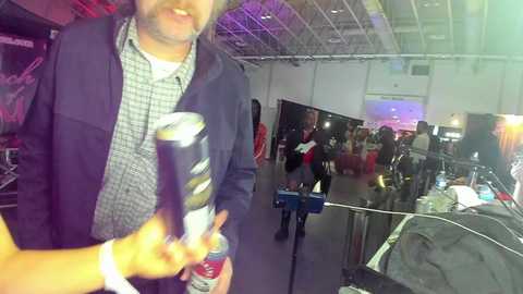 Media: Video of a bearded man in a plaid shirt and navy jacket holding a beer can, inside a dimly lit, cluttered convention center with numerous people and booths in the background.