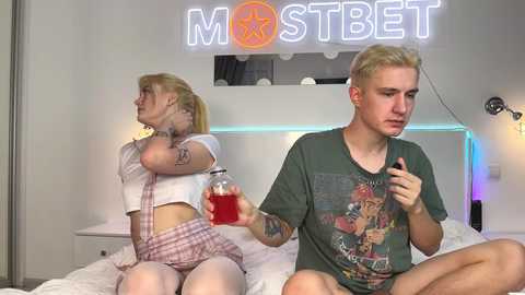 Media: Video of a blonde woman in plaid shorts and crop top, holding a red drink, and a young man with short hair, wearing a graphic tee, sitting on a bed with white sheets in a modern, minimalist room.