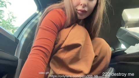 Media: Video of a young woman with long brown hair, wearing a red long-sleeve top and brown pants, sitting in a car, lips slightly parted, URL watermark visible.