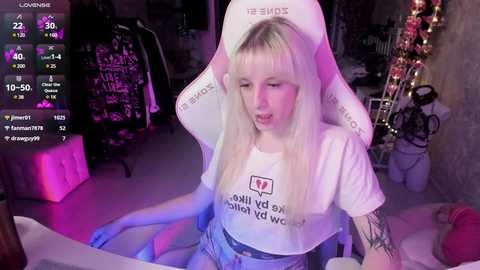 Media: Video of a young, pale-skinned, blonde woman with straight hair, wearing a white crop top with \"FUCK YOU\" text and blue jeans, sitting in a white gaming chair, in a dimly lit room with a neon sign and gaming setup.