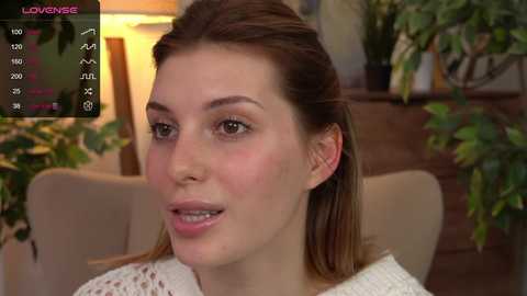 Media: Video of a fair-skinned woman with straight, light brown hair, wearing a white knitted sweater, smiling, in a cozy indoor setting with plants and wooden furniture.