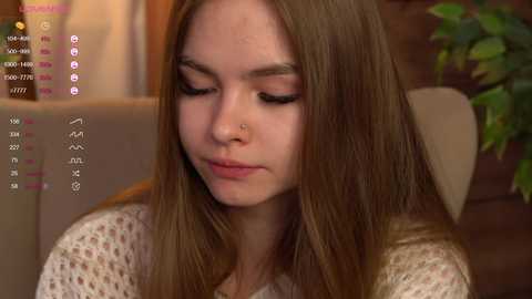 Media: Video of a young Caucasian woman with long brown hair, wearing a white knit sweater, sitting indoors. Background includes a blurred couch and green potted plant.