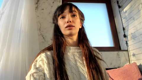 Media: Video of a young Asian woman with long brown hair and fair skin, wearing a cream-colored cable-knit sweater, standing in a dimly lit room with a window and white curtains.