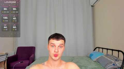 Media: Video of a young, shirtless man with fair skin, short dark hair, and a playful expression, lying on a bed with light green sheets. Background includes a purple chair, white curtains, and an air conditioning unit.