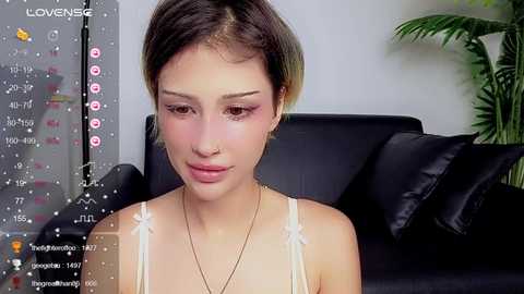 Media: Video of a young woman with short, dark brown hair and fair skin, wearing a white spaghetti-strap top, sitting indoors on a black leather couch. A plant is in the background.