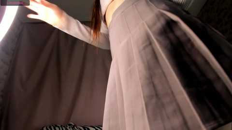 Media: Video of a woman with long, straight brown hair wearing a grey pleated skirt, standing in a dimly lit room. The background features zebra-patterned fabric and a blurred, dark wall.