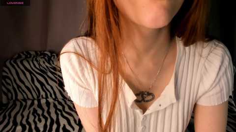 Media: Video of a young woman with long, straight red hair, fair skin, and a white ribbed top, revealing a necklace with a heart pendant. Background includes a zebra-print blanket.