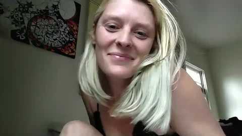 Media: Video of a smiling, fair-skinned woman with platinum blonde hair, wearing a black tank top, in a dimly lit room with abstract wall art.