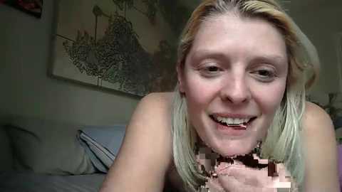 Media: A close-up video of a smiling blonde woman with light skin, topless, with a pixelated penis in her mouth, set in a dimly lit room with a framed art piece in the background.