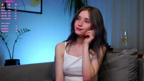 Media: Video of a fair-skinned, young woman with long, straight brown hair, wearing a white spaghetti-strap top, sitting on a gray sofa. Background includes a potted plant, framed artwork, and a glass of wine.