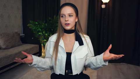 Media: Video of a Caucasian woman with long, straight, light brown hair, wearing a white jacket over a black top and a black choker. She gestures with open arms, standing indoors with a beige couch, dark curtains, and a plant in the background.