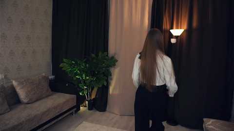 Media: Video of a woman with long hair in a white striped blouse and black pants standing in front of a beige curtain, dimly lit room with a potted plant and a cushioned beige sofa.