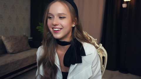 Media: Video of a smiling young girl with light skin and long, wavy brown hair. She wears a black beret, black choker, and a beige blazer with a black bow tie. Background features a beige couch with cushions, a green plant, and a dimly lit room.