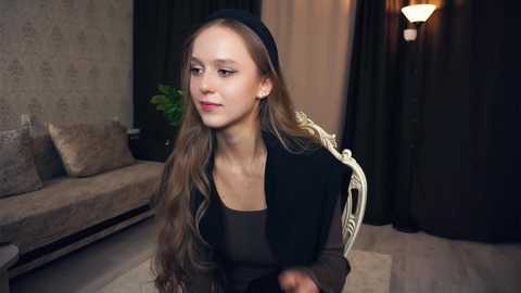 Media: Video of a young woman with long, wavy brown hair, wearing a black beret, black top, and black vest, seated in a modern living room with beige and gray decor, dark curtains, and a green plant.