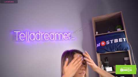 Media: Video of a woman with light skin and long brown hair, wearing a purple top, covering her eyes. Neon \"Telladreamer\" sign on a grey wall, behind a wooden shelf with a blue \"MSTREET\" hat.