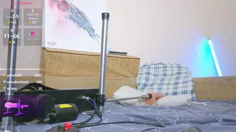 Media: A video of a bedroom with a messy bed featuring a white pillow, a blue and white checkered blanket, and a black vibrator with a red base on the sheets. A blue light illuminates the room.