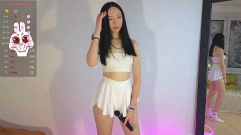 Media: Video of a young East Asian woman with long black hair, fair skin, and medium breasts, dressed in a white crop top and skirt, posing in a modern room with a mirror.