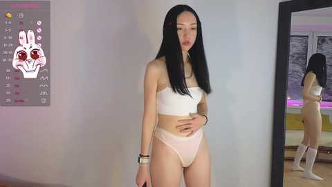 Media: Video of an East Asian woman with long black hair, fair skin, and medium-sized breasts, wearing a white bandeau top and matching panties, standing in a modern, minimalist room with a mirror and abstract wall art.