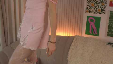 Media: Video of a person in a pink lace dress, standing beside a bed with beige curtains and a green pillow. Background features a colorful, abstract art piece.