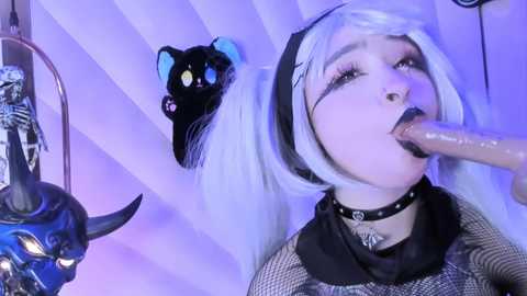 Media: Video of a young woman with pale skin, white hair in twin pigtails, wearing a black choker and fishnet top, performing oral sex on a large, erect penis, set in a room with a black cat mask and a skull on the wall.