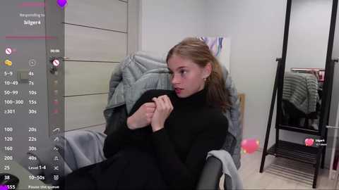 Media: Video of a young woman with light skin, long brown hair, and fair complexion, sitting on a chair, wearing a black turtleneck, clutching a gray blanket. Background includes a mirror, pink balloons, and a white wall.