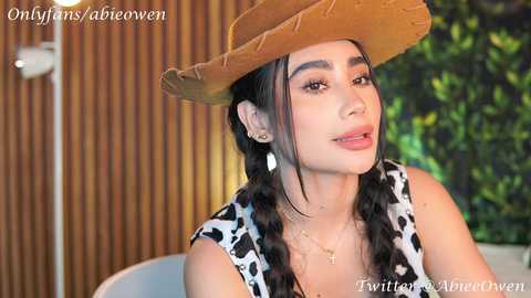 Media: Video of a young woman with long, dark hair in braids, wearing a cowprint dress and straw hat, against a wooden backdrop.