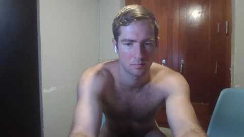 Media: Video of a shirtless, muscular, Caucasian man with short brown hair, looking down, in a dimly lit room with a wooden door and blue chair.