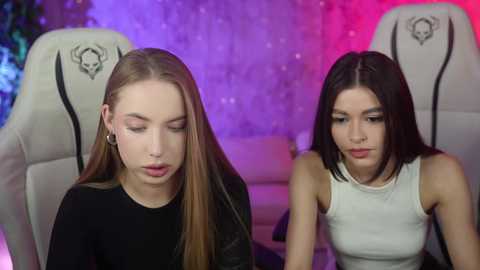 Media: Video of two young women with light skin and straight hair in a gaming setup. One wears a black top, the other a white tank top. Background features a purple and red LED light, white gaming chairs, and a white wall.