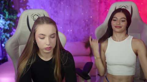 Media: Video of two young women with long hair, one in a black top, the other in a white crop top, seated on gaming chairs in a colorful, dimly lit room with purple and pink lights.