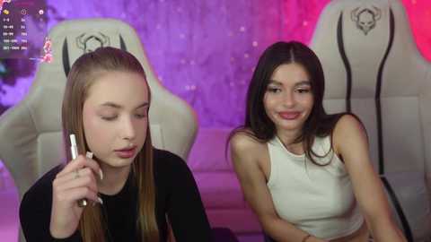 Media: Video of two young women with light skin, one with long blonde hair in a black top, the other with long dark hair in a white crop top, sitting in white gaming chairs with purple and red LED lights in the background.