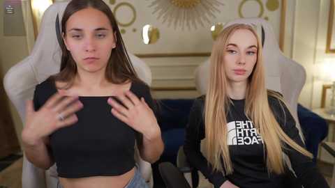 Media: Video of two young women in a gaming setup. One, a brunette, wears a black crop top, the other, a blonde, wears a black shirt with white text.