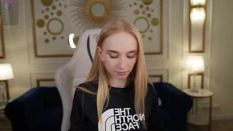 Media: Video of a blonde woman in a black sweatshirt with white text, standing in a well-lit, elegantly decorated room with gold accents, a plush blue couch, and a lamp.