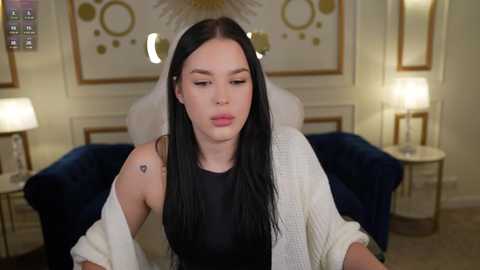 Media: Video of a pale-skinned woman with long black hair, wearing a black sleeveless top, sitting on a plush navy-blue sofa in a well-lit, elegantly decorated room with framed art and lamps.