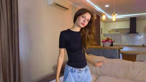 Media: Video of a young woman with long brown hair, wearing a black t-shirt and blue jeans, standing in a modern, well-lit living room with a kitchen visible in the background.