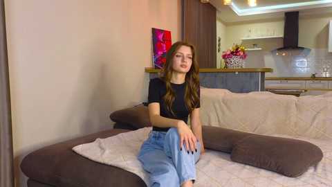 Media: Video of a young Caucasian woman with long brown hair, wearing a black top and light blue jeans, sitting on a beige sectional sofa in a modern, warmly lit living room with a kitchen visible in the background.