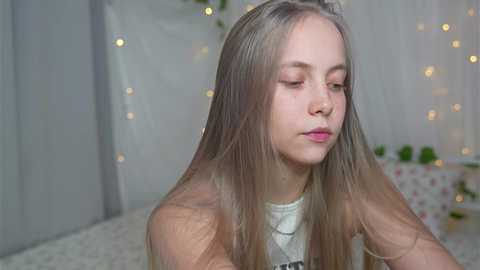 Media: Video of a young girl with long, straight blonde hair, fair skin, and closed eyes, wearing a white top, in a softly lit, white bedroom with string lights and a floral patterned table in the background.