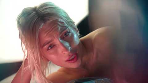 Media: Video of a close-up, intimate moment between a blonde woman with wet hair and a man with visible, muscular shoulders. They are naked, engaging in oral sex. The background is blurred, with soft, ambient lighting.