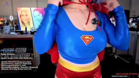 Media: Video of a woman in a blue, long-sleeve, shiny, Superman-themed crop top, yellow belt, and red shorts, lifting her shirt, revealing a tattoo. Background shows a cluttered room with a desk, computer, and a large screen displaying a woman's face.