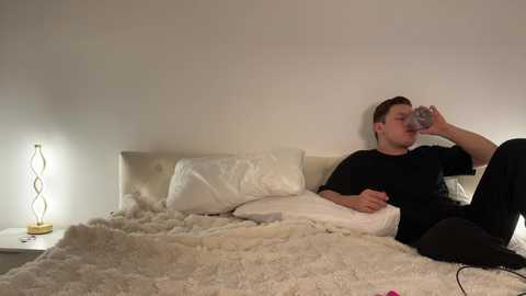 Media: Video of a young man in a black T-shirt, drinking water from a glass, lying on a white bed with a white duvet and fluffy blanket in a minimalist, beige-walled bedroom.