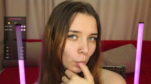 Media: Video of a young Caucasian woman with long brown hair, blue eyes, and a nose ring, playfully biting her finger in front of a red couch, overlaid with virtual reality elements.