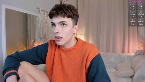 Media: Video of a young, fair-skinned, short-haired man with a slim build, wearing a blue and orange sweater, sitting on a light-colored couch. Background includes a beige curtain and a partially visible screen with gaming stats.