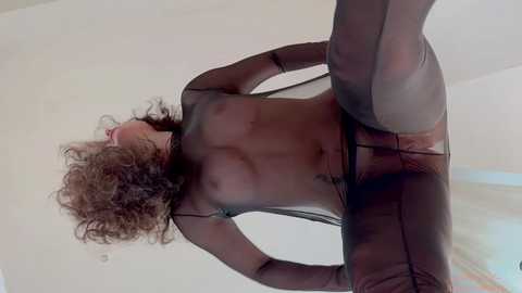 Media: Video of a woman with curly brown hair, wearing a sheer black bodysuit, posing provocatively against a plain white background, emphasizing her slender physique and medium-sized breasts.