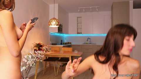 Media: Video of a modern kitchen with two nude women: one holding a phone, the other in the foreground, blurred.