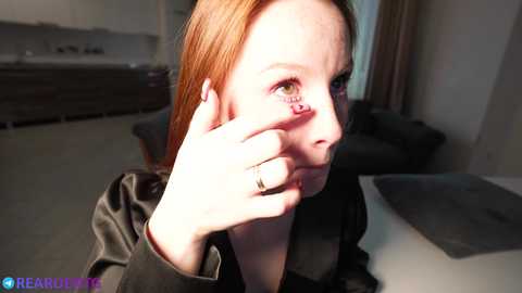 Media: Video of a fair-skinned woman with long, straight red hair, wearing a black shirt, touching her face with a gold ring on her finger. Background shows a modern living room with beige carpet and gray furniture.