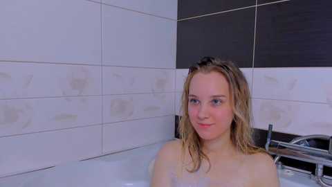 Media: Video of a young Caucasian woman with wet, blonde hair, fair skin, and blue eyes, sitting in a white bathtub with black and white tiled walls.