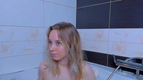 Media: Video of a young, light-skinned, blonde woman with shoulder-length hair, nude, sitting in a modern, tiled bathroom shower, looking contemplative.