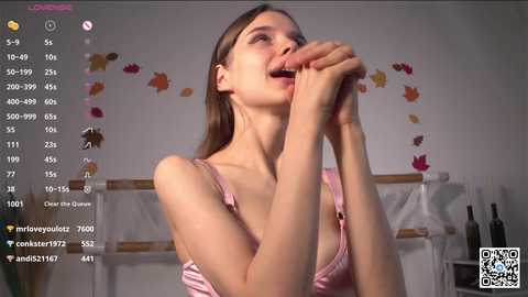 Media: A video of a young woman with long brown hair, wearing a pink spaghetti-strap top, licking her fingers in a cozy room with autumn-themed decor.