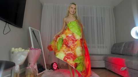 Media: A video of a blonde woman in a vibrant, flowing dress, standing in a modern living room with white walls, a large window, and a grey sofa.