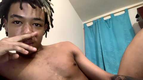 Media: Video of a shirtless Black man with dreadlocks, a nose piercing, and a facial scar, sitting in a bathroom with blue shower curtain.