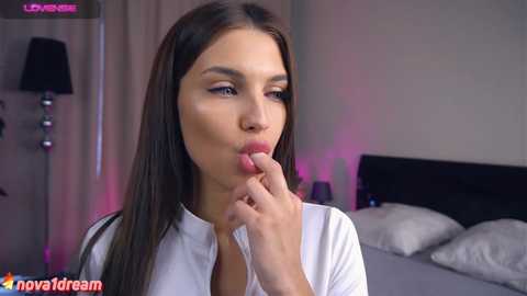 Media: Video of a young woman with straight brown hair and fair skin, wearing a white blouse, seductively licking her fingers in a bedroom with a black lamp and grey bedding.